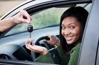 A hand handing car keys to a lady looking at the camera sitting inside a car with the window down